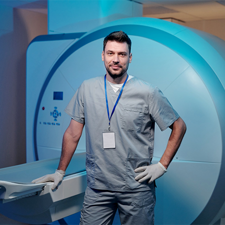 A male MR technologist in the MRI room. The MRI sits behind him, blue-teal light shining on it.