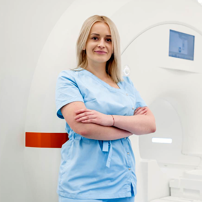 An MR personnel by an MRI machine, arms cross and looking into the camera.