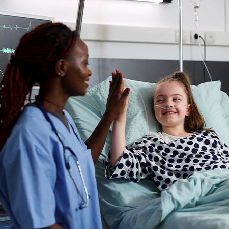 A female nurse giving a high five to a young, ill pediatric patient in the patient's room.