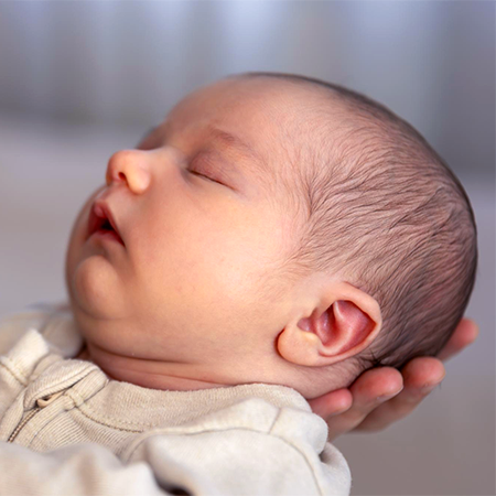 A sleeping baby in a tan onesie being held up.