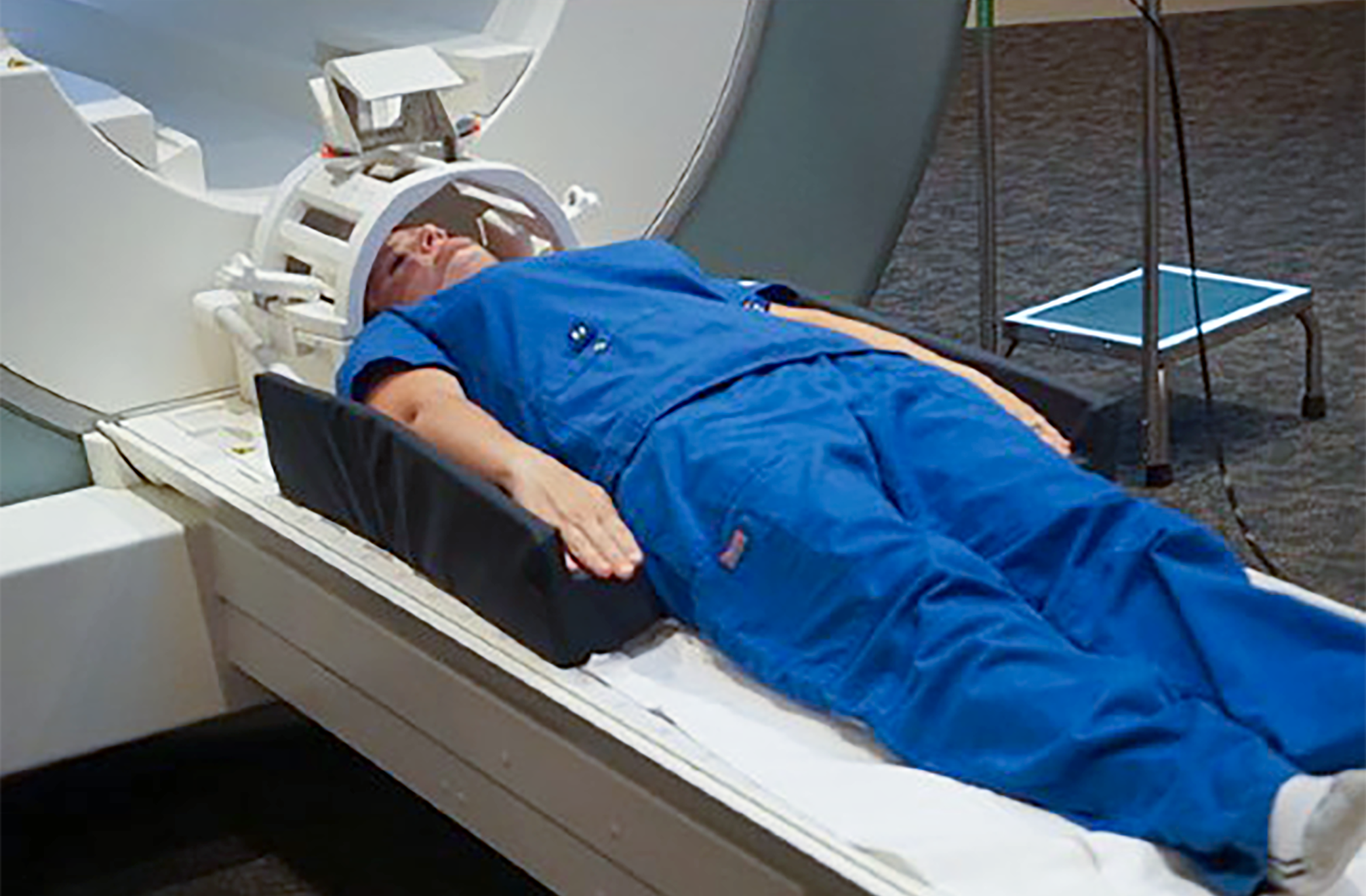 A patient with a head coil and dark blue scrubs is laying on the scan table. Arms are cupped in L-shaped foam positioning aids. Image links to product page.