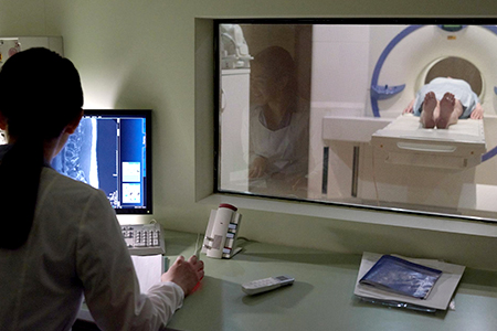 An MR personnel is in the MRI control room studying a monitor. A patient is on the scan table.