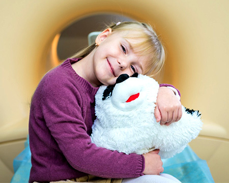A young female MRI pediatric patient smiling and hugging a large black and white stuff animal.